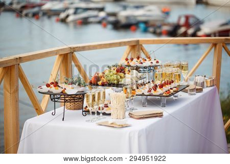 On The Table With A White Cloth There Are Glasses Of Wine, Trays Of Grapes, Peaches, Cherries And Ta