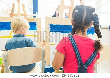 A Group Of Preschool Students And Young Teacher In Drawing Class Paint In Gouache. Mixed Race School