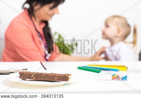 On The Table There Is A Plate With Chocolate, In The Background The Doctor Examines A Little Girl An
