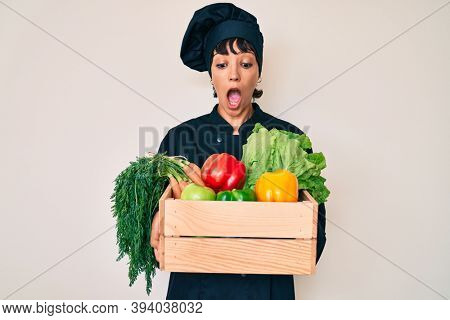 Beautiful brunettte woman chef holding fresh veggetables afraid and shocked with surprise and amazed expression, fear and excited face. 