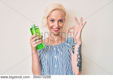 Young blonde woman with tattoo holding aloe vera doing ok sign with fingers, smiling friendly gesturing excellent symbol 