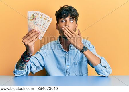 Young hispanic man holding united kingdom 10 pounds banknotes sitting on the table covering mouth with hand, shocked and afraid for mistake. surprised expression 