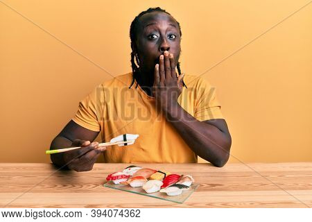 Handsome young black man eating sushi sitting on the table covering mouth with hand, shocked and afraid for mistake. surprised expression 