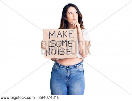 Beautiful young brunette woman holding make some noise banner serious face thinking about question with hand on chin, thoughtful about confusing idea 