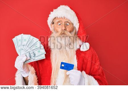 Old senior man wearing santa claus costume holding dollars and credit card making fish face with mouth and squinting eyes, crazy and comical. 