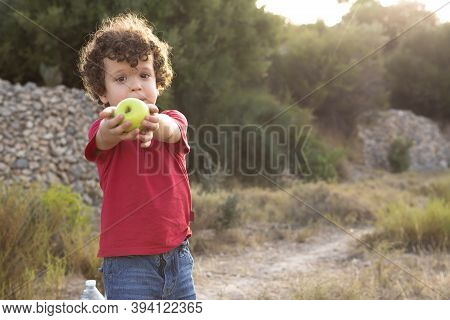 Beautiful Curly-haired Boy In The Field And Dressed In Red, Offering A Yellow Apple 