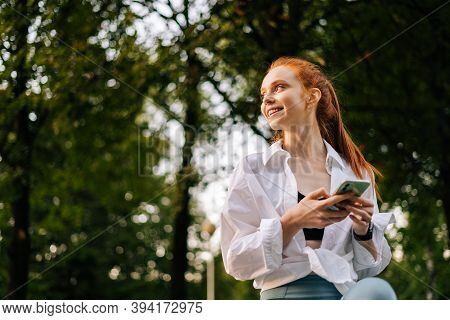 Low-angle Shot Of Cheerful Young Woman Browsing Internet On Smartphone, Looking Away. Portrait Of Fe