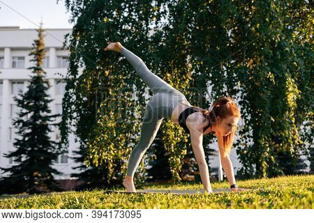 Low-angle Shot Of Young Woman Practicing Yoga In Down Facing Dog Pose On One Leg, City Park At Sunny