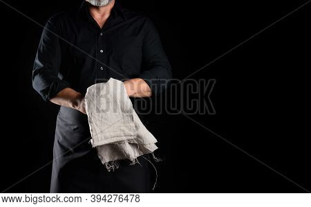 Male Chef In A Black Apron And Shirt Holds A Gray Linen Napkin On A Black Background, Place For An I
