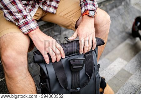 Young Caucasian Man Opens A Black Roll Top Backpack While Sitting On A Bench Near An Electric Scoote