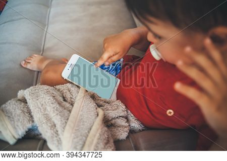 Caucasian Small Boy Is Sitting In Bed And Holding A Phone While Looking At The Screen Covered With A