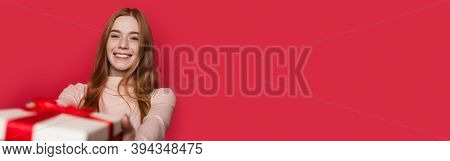 Adorable Woman With Freckles And Red Hair Is Smiling At Camera Receiving A Present On A Red Studio W