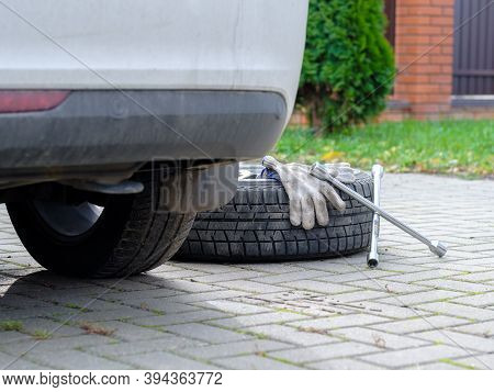 A Wrench And Work Gloves Lie On A Winter Car Wheel Lying On The Ground. There Is A Car Nearby. The C