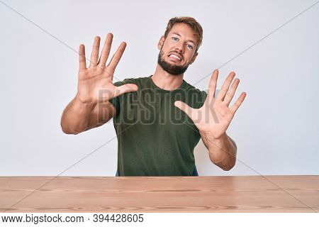 Young caucasian man wearing casual clothes sitting on the table afraid and terrified with fear expression stop gesture with hands, shouting in shock. panic concept. 