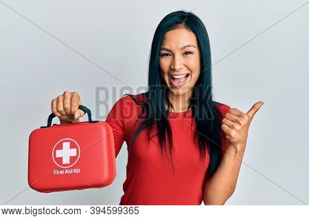 Beautiful hispanic woman holding first aid kit pointing thumb up to the side smiling happy with open mouth 