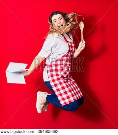 Young beautiful blonde cook woman wearing apron smiling happy. Jumping with smile on face holding recipe book and spoon over isolated red background