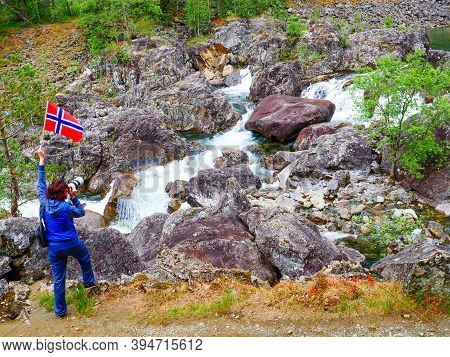 Tourist Woman With Norwegian Flag And Camera Walking Trough Mountains Path In Allmannajuvet Area Sau