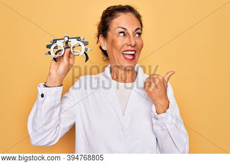 Middle age senior oculist woman holding optometrical glasses for vision correction pointing and showing with thumb up to the side with happy face smiling