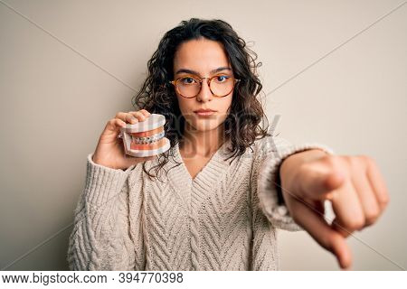 Young beautiful woman with curly hair holding plastic teeth with dental baces pointing with finger to the camera and to you, hand sign, positive and confident gesture from the front