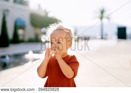 Toddler In A Terracotta Dress Is Eating Pirogi By The Fountain In The Park On A Sunny Day