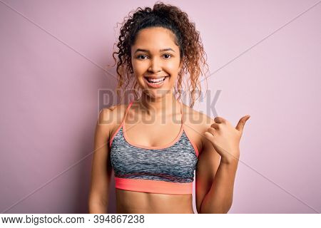 Beautiful sportswoman with curly hair doing sport wearing sportswear over pink background smiling with happy face looking and pointing to the side with thumb up.