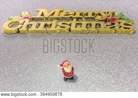 Cute Tiny Figurine Of Santa Claus On A Glitter Silver Snow Background With The Golden Words Merry Ch