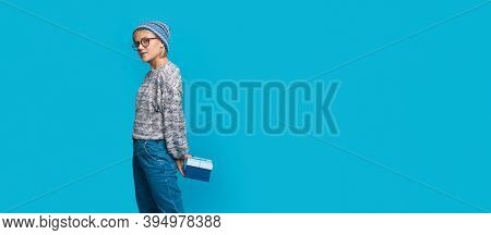 Adorable Woman With Glasses And Hat Is Holding A Present At Her Back While Looking At Camera On A Bl