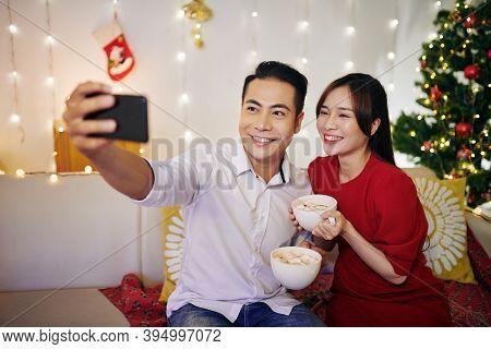 Cheerful Beautiful Young Asian Couple Taking Selfie With Cups Of Hot Cocoa In Hands On Winter Evenin