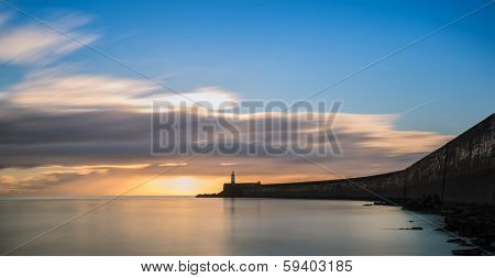 Beautiful Vibrant Sunrise Sky Over Calm Water Ocean With Lighthouse And Harbor Wall