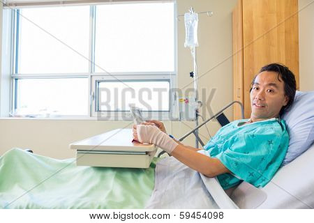 Portrait of male patient holding digital tablet while reclining on bed in hospital
