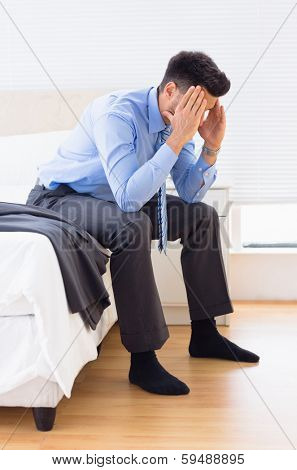 Hungover businessman sitting at edge of bed at home in bedroom
