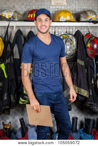 Portrait of happy firefighter holding clipboard against uniforms hanging at fire station