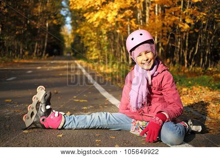 Happy girl in a protective helmet sits outstretched leg in the rollers on the asphalt in the autumn park