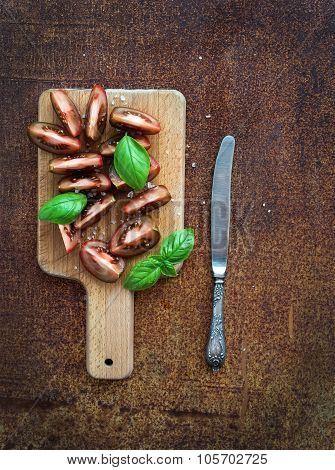 Ripe kumato tomatoes, basil leaves and knife on a rustic wooden chopping board over grunge rusty met