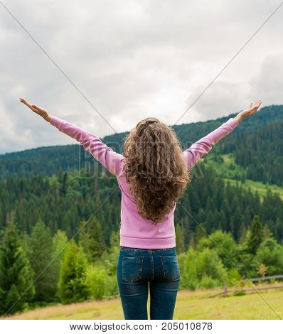 Curly woman happy smiling joyful with arms up dancing on top peak of mountains in summer during holidays. Beautiful young cheerful female model standing back to camera.Young woman outdoors.Freedom concept.