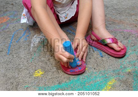 Little girl creatively paints her toe nails with blue sidewalk chalk.