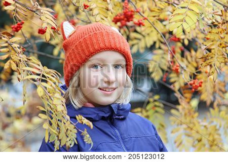 Cheerful girl smiling, the child is dressed in a funny knitted Cheerful kid girl smiling, the child is dressed in a funny knitted warm hat with ears, looks like a fox. Autumn, outdoors portrait with yellow leaves and rowan berries.