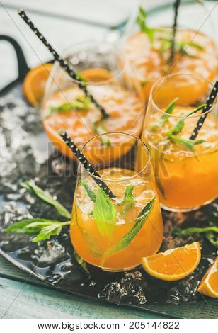 Refreshing cold alcoholic summer citrus cocktail with orange, peppermint and crushed ice in stemless glasses on dark tray over light wooden background, selective focus, vertical composition