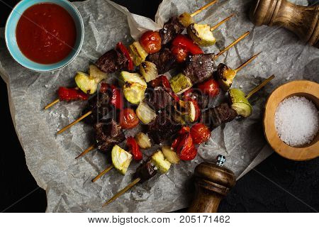 Grilled meat and vegetables cabobs with tomato sauce and sea salt over black stone table. Top view