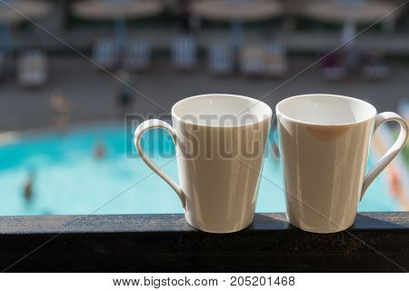 Two White Mugs Stand On The Balcony In The Background Of The Hotel, Where The Buildings And The Pool