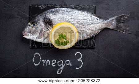 Raw dorada fish laying on black slate and written word omega 3 by white chalk.