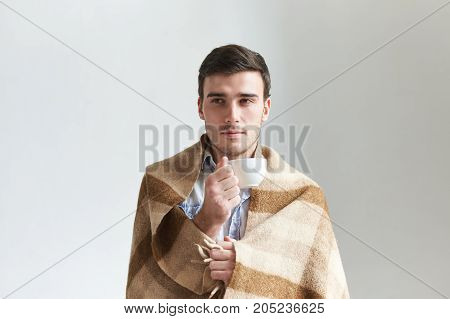 Attractive young male in checkered plaid holding cup of hot coffee warming himself after long walk on cold autumn day. Brunette man wrapped in warm wool blanket enjoying herbal tea at home