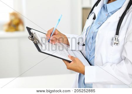 Close-up of a female doctor filling  up medical form at clipboard while standing straight in hospital.