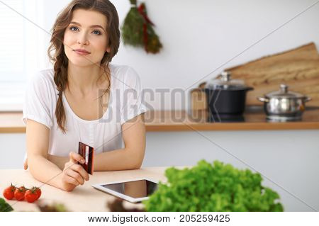 Young  woman looking for a new recipe for  cooking in a kitchen. Housewife is making online shopping by tablet computer and credit card.