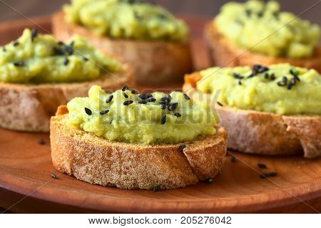 Bread slices with green pea and parsley spread garnished with black sesame seeds served on wooden plate photographed with natural light (Selective Focus Focus on the front of the spread on the first bread)