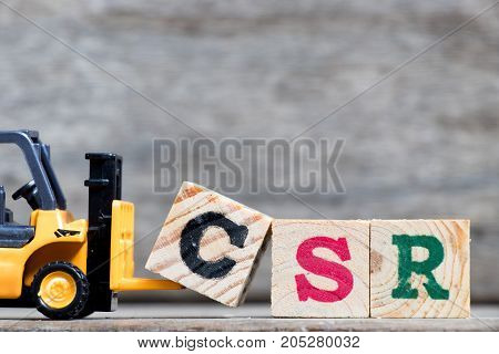Yellow plastic forklift hold letter C to complete word CSR (Abbreviation from corporate social responsibility) on wood background