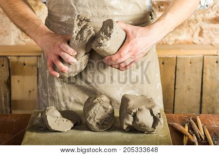 potter, tools, ceramics art concept - front view on hands of young master holding the large pieces of fireclay, male dressed in an apron, a ceramist with raw materials on table