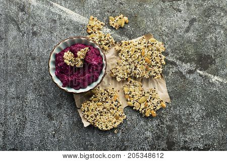 Crispy slices of homemade flat bread without gluten from the seeds of quinoa, pumpkin, sunflower, sesame with the addition of corn flour, water and vegetable oil on a gray background with hummus from the baked organic beetroot. Top View