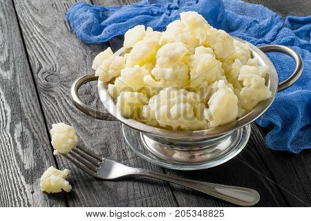 Blanched cauliflower in colander on gray wooden background. White cauliflower inflorescences in metal colander fork and blue gauze napkin on table. Preparing for diet dinner