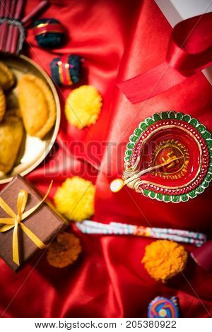stock photo showing Diwali still life with Diya or oil Lamp, gift boxes, sweets and fire crackers with flowers arranged over red satin cloth with bokeh